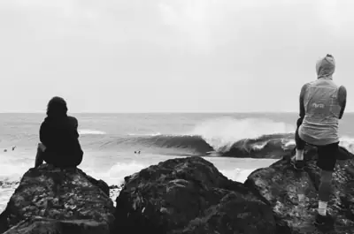 Two people sitting on rocks watching breaking surf, in black and white