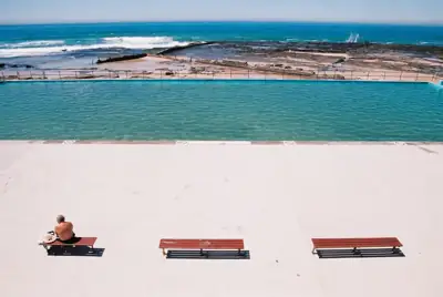 Person sunbathing beside an ocean pool with red benches along the promenade