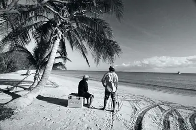 Two people with a bodyboard beneath palm trees beside a beach, in black and white