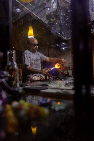 Glassblower shaping glowing orange glass at a workbench, seen through workshop reflections